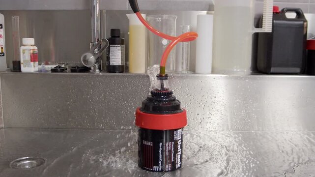Film developing tank is rinsed with running water in a professional darkroom laboratory. Surrounding measuring jugs, bottles, and chemistry highlight the analog black-and-white film process.