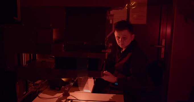 Photo lab worker uses an analog photographic enlarger under red safelight in a darkroom. The process enables precise, manual creation of silver-gelatin prints from film negatives.