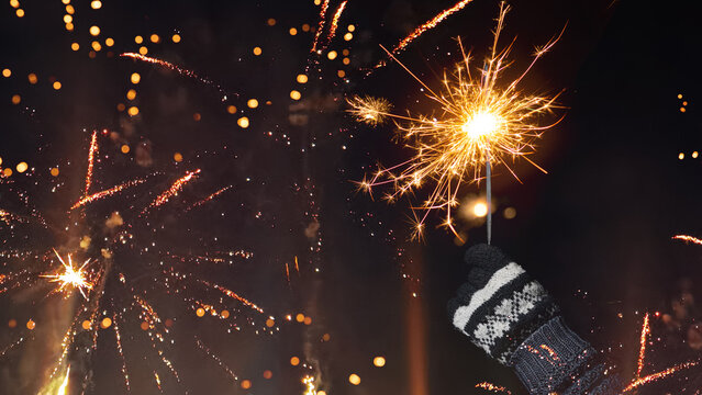 Happy new year, new years's eve holiday celebration greeting card - woman's hand with glowing sparkler on dark night sky with firework - Powered by Adobe