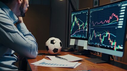Man analyzes stock market trends while sitting at a desk with soccer ball and two computer screens displaying data - Powered by Adobe