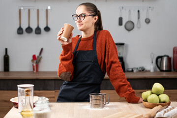 Woman housewife in apron and eyeglasses drinking takeaway delivery paper cup coffee while cooking food in the kitchen at home