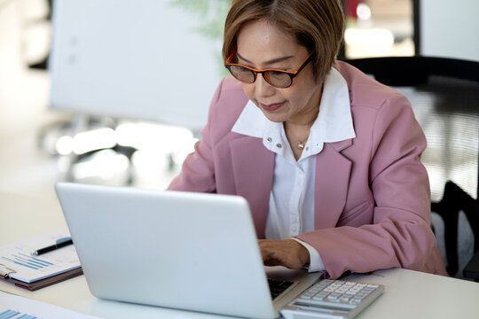Asian businesswoman professional adult working laptop computer office calculating financial data managing strategy corporate meeting room executive typing reports analyzing graphs focused diligent