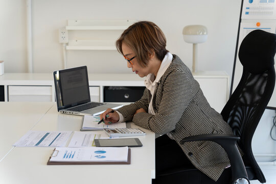 Adult Asian businesswoman analyzing financial reports charts documents planning corporate strategy at modern home office workspace