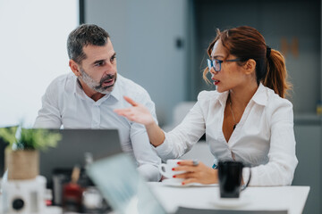 Two colleagues in white shirts collaborate at a sleek office desk, animatedly discussing ideas as...