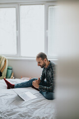 Obraz premium A man wearing a plaid shirt and red socks sits on a bed working on a laptop in a casual home environment. He displays focus and engagement during this remote working moment.