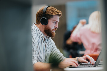 A bearded man wearing large over-ear headphones concentrates on his laptop at a modern desk. The...