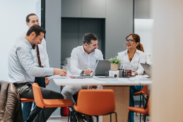 A diverse group of coworkers gathers around a table with laptops in a bright, modern office, sharing ideas and enjoying a friendly, productive moment.
