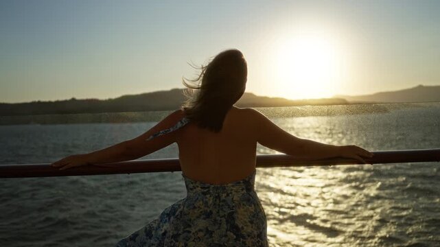 Woman with bare back leaning on ship rail at deck watching the golden sunset, dressed in floral halter dress with hair blowing in wind; quiet serenity.