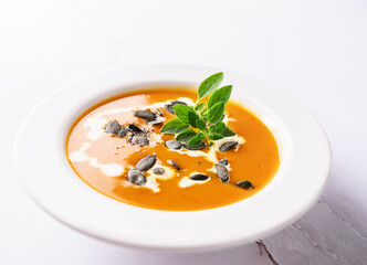 Hot spicy pumpkin soup with seeds, oregano leaves and bread in white ceramic plate on light marble background close up. Healthy vegan food concept.