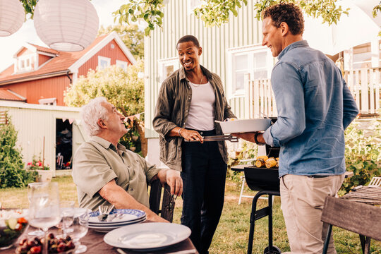 Male friends talking with senior man while barbecuing in back yard at social gathering party