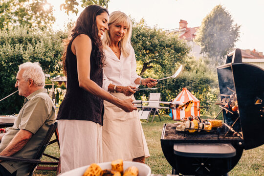Woman grilling vegetable skewers on barbecue with mother in back yard