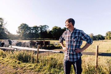 Farmer looking away and standing with arms akimbo near fence at cattle farm