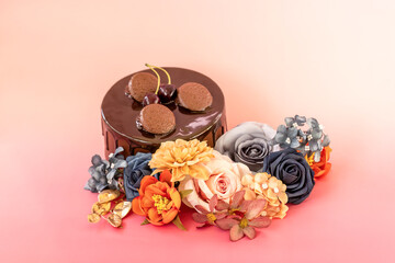 Elegant chocolate cake decorated with cherries and small pastries, surrounded by a colorful floral arrangement of artificial flowers in blue, orange, and pastel tones on a pink background