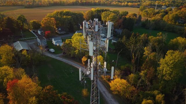 Aerial view of a telecommunications tower surrounded by autumn forest and rural houses in warm sunset light.