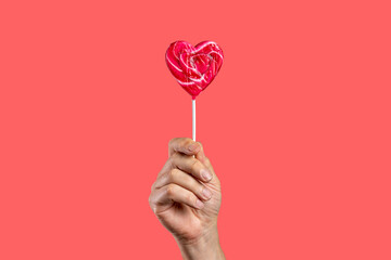 Hand holding heart-shaped lollipop on pink background. Valentine's Day concept. Man holding red...