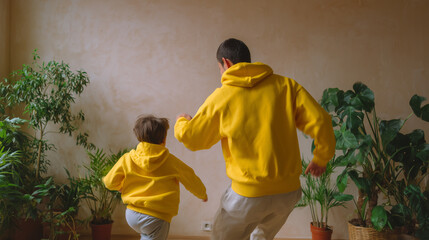 Joyful father and son in matching yellow hoodies dancing or playing at home, surrounded by green potted plants. Family fun and bonding.