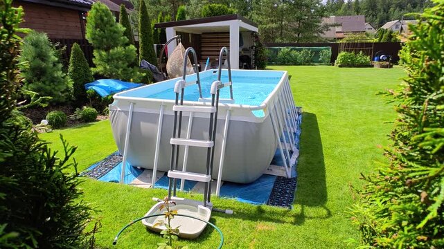 On the mowed grassy lawn stands a gray PVC rectangular frame pool with a ladder. The water in it is filtered. Nearby is a wooden gazebo, swings, thujas, metal fence, bushes and trees. Sunny summer