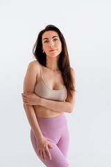 Fototapeta premium Sports woman standing in activewear with abs muscles, looking at camera isolated on white background. Caucasian 30s fitness girl wears beige top, pink leggings. Happy fit smiling brunette, blue eyes.