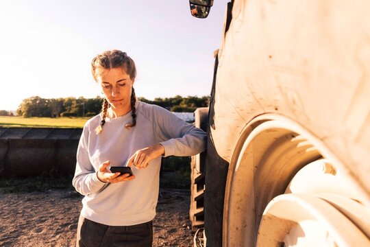 Female farmer with braided hair scrolling smart phone while leaning on tractor   in farm
