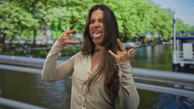 Woman showing rock hand gesture and tongue out by canal railing on amsterdam street during daytime; playful defiance.