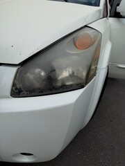 Close-up of a severely oxidized, yellowed, and peeling car headlight on a white vehicle, illustrating the need for restoration, repair, or replacement due to damage and wear.