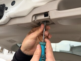 A mechanic's hands using a flathead screwdriver to carefully remove a plastic trim fastener from the interior panel of a car's trunk or tailgate, demonstrating vehicle disassembly.