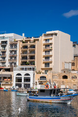 Midday sun lights pastel apartment blocks with balconies in Sliema, Malta as luzzus bob on calm reflective water, limestone facades and arches line the compact bay.