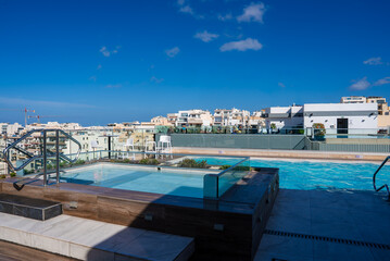 A rooftop infinity pool and hot tub sit above Mellieha, Malta, with cream low rise buildings, glass railings, and wood edges in bright midday light and calm water.