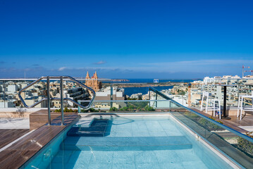 A rooftop plunge pool with glass balustrades overlooks Mellieha, Malta. Mellieha Parish Church rises above town, with Mellieha Bay and the channel to Comino and Gozo visible.