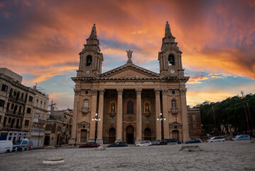 Obraz premium A Baroque church with twin bell towers stands in a Valletta square at sunset, facade lit with warm bulbs, clocks on towers, statue on pediment, cars and townhouses nearby.
