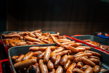 Close up trays piled with grilled sausages under warm indoor light. Browned casings glisten, shallow depth of field blurs background, buffet style abundance is clear.