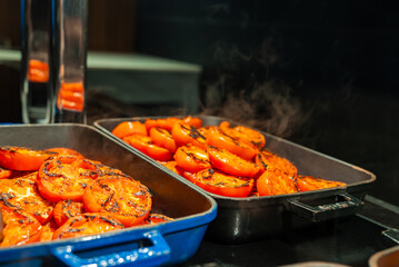 Two oven trays hold steaming char grilled tomato halves on a stovetop in Mellieha, Malta. Warm light highlights blistered skins, herbs, oil, and rising steam.