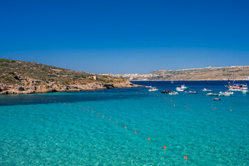 Obraz premium Turquoise water fills the Blue Lagoon on Comino, Malta. Boats anchor near rocky cliffs as swimmers drift along a buoy line. Bright noon light and wide angle highlight clarity.