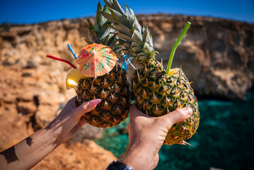 Two hands clink pineapple drinks with straws and a small umbrella near the Blue Lagoon on Comino, Malta, bright daylight, saturated colors, shallow depth of field