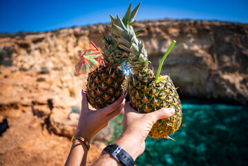 Two hands raise hollowed pineapples with straws and umbrellas, toasting by a rugged limestone cliff and turquoise water at Blue Lagoon, Malta, in bright midday light.