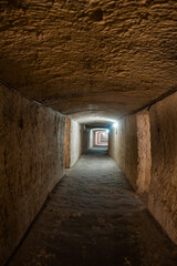 Obraz premium A stone lined WW2 Shelter tunnel in Mellieha, Malta recedes with low arches and cool white lights, empty and textured, centered perspective, soft ambient light, daytime indoor setting.