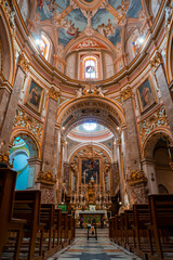 Obraz premium Baroque church interior in Mdina, Malta shows marble columns, gilded stucco, high altar, wooden pews, arches, paintings, frescoed ceilings, and oculi windows in soft light.