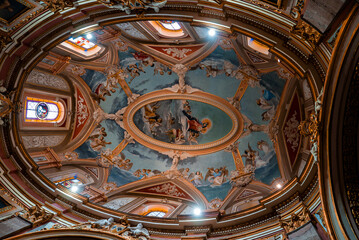 Obraz premium A Baroque dome in Mdina, Malta is seen from below, with pastel blue frescoes of angels and saints, gilded stucco, stained glass windows, and warm light in a wide angle view.
