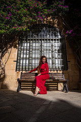 A woman in a long red dress sits on a bench by an iron barred arched window in Mdina, Malta....