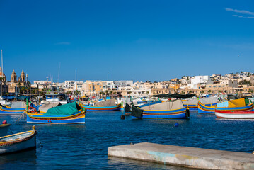 Obraz premium Daytime view of Marsaxlokk harbor, Malta, with luzzu boats bobbing on deep blue water, tarps on decks, honey colored town, and the Parish Church towers and dome.