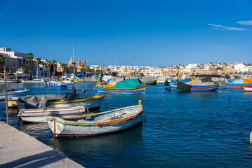 Brightly painted luzzu boats bob in Marsaxlokk, Malta, as reflections dance on deep blue water. The church dome and twin bell facade rise beyond limestone quays on a clear day.