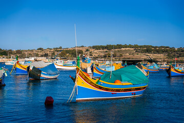 Obraz premium Traditional Maltese luzzu boats float in Marsaxlokk, Malta, their vivid colors reflecting on deep blue water. Afternoon light reveals canopies, nets, and terraced hills.