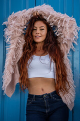 A young woman lifts a blush pink cardigan over her head at a vivid blue door in Marsaxlokk, Malta....