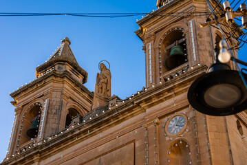 Close up of the Church of Our Lady of Pompei in Marsaxlokk, twin bell towers with string lights, clock, green bell, central saint statue, warm late afternoon light.