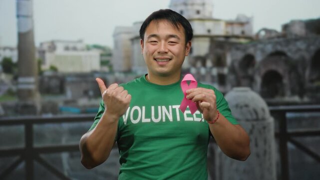 Young man volunteer holding pink ribbon in vatican san pedro square promoting breast cancer awareness with positive expression outdoors in italy street scene.