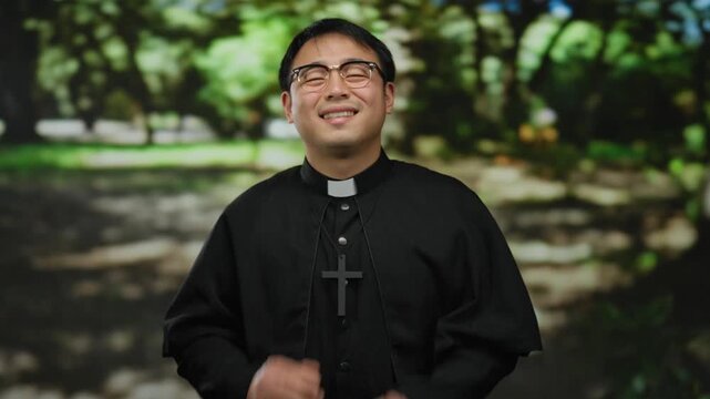 Asian man smiling in a park wearing a clerical collar and cross, surrounded by greenery and sunlight, showcasing joy and faith in an outdoor setting.