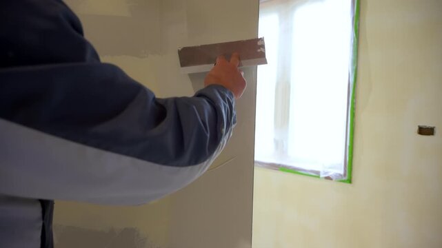 Construction worker spreading joint compound on drywall wall corner using a metal taping knife. Wall smoothed and finished as part of a home renovation or remodeling project