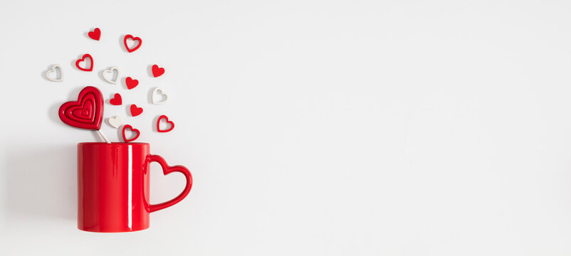 Valentine’s Day composition with red mug, heart-shaped handle, and red lollipop on white background. Minimal love concept with small red and white hearts, romantic flat lay with copy space