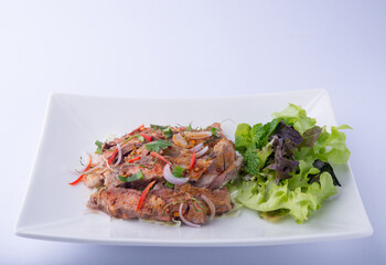 Thai food. Grilled Pork Neck Salad, pork cut into long strips, garnished with chilies, shallots, cilantro, and salad greens and mint leaves on a white plate, white background.