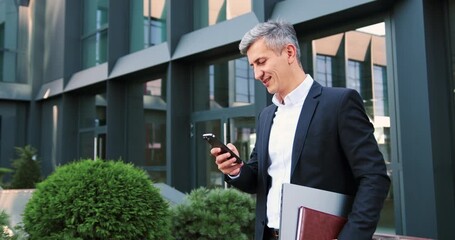 Man going outdoor with mobile phone in his hands. Middle aged businessman walking using smartphone outdoors in city near office building. - Powered by Adobe